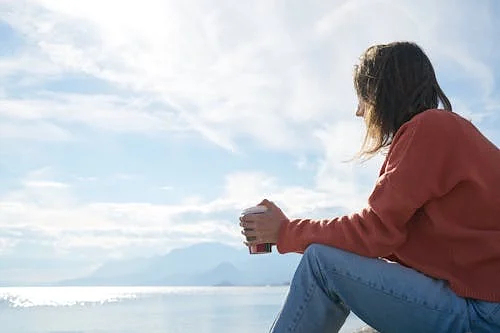 Free A woman in casual attire enjoys a hot drink while gazing at the sea, epitomizing relaxation. Stock Photo